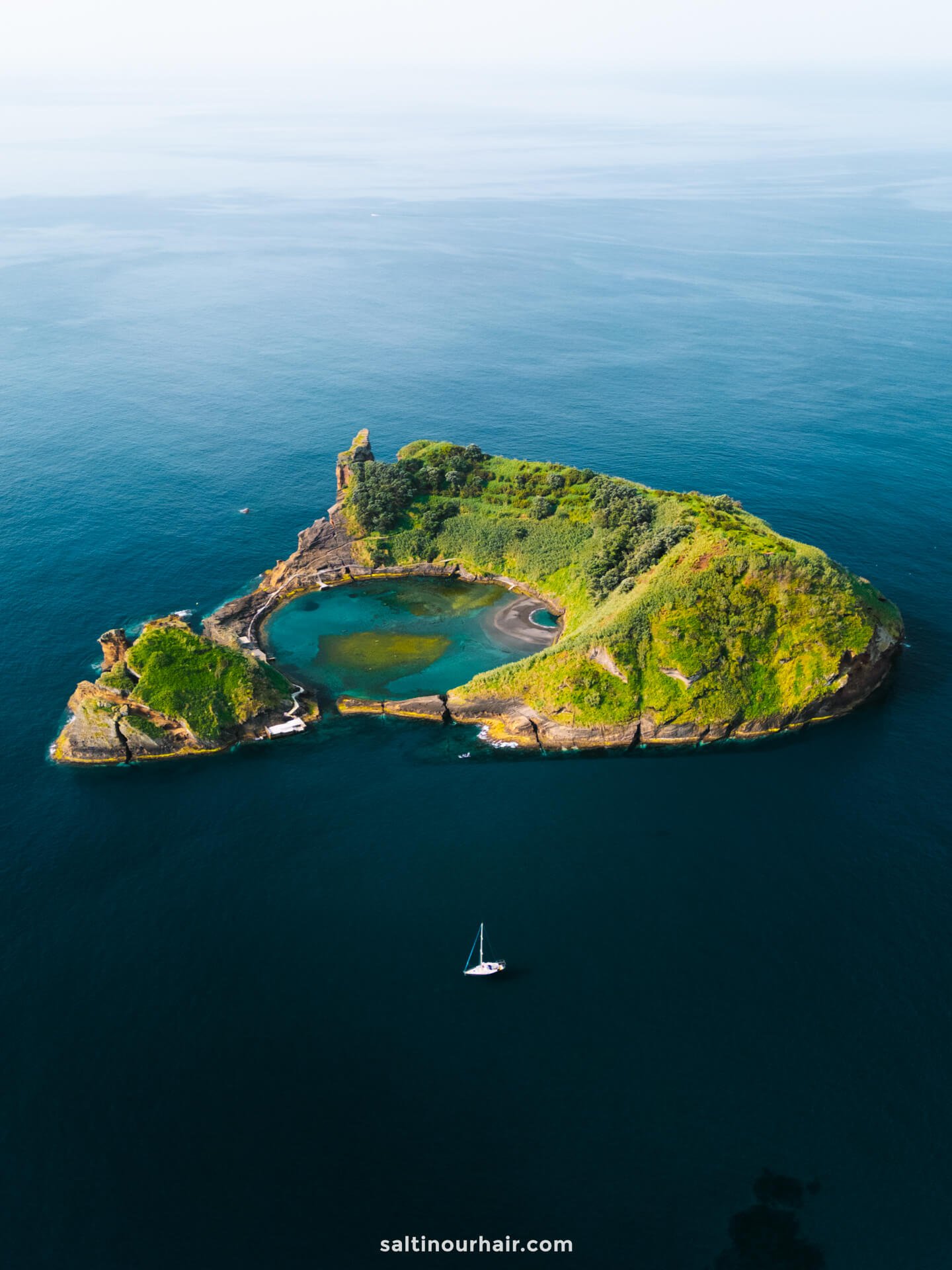 A small, green, rocky island with a central lagoon sits in calm blue ocean waters; a single sailboat floats nearbyâ€”evoking the serene beauty and tranquil escapes found among the best things to do in SÃ£o Miguel, Azores.
