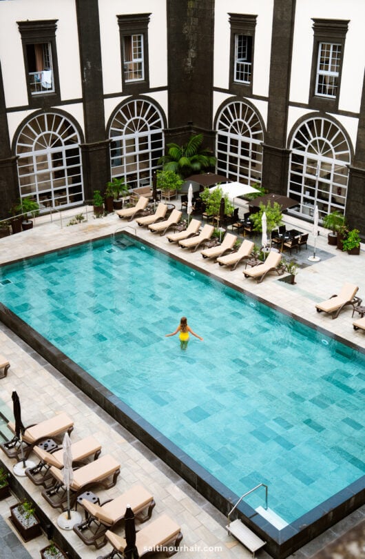 A person in a yellow swimsuit stands in the center of a rectangular outdoor pool—one of the relaxing things to do in São Miguel—surrounded by lounge chairs and a building with arched windows.