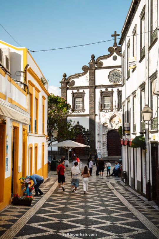 A cobblestone street with people walking, lined with colorful buildings, leads to a white church reminiscent of Ponta Delgada, Azores, featuring dark stone accents and a large rose window.