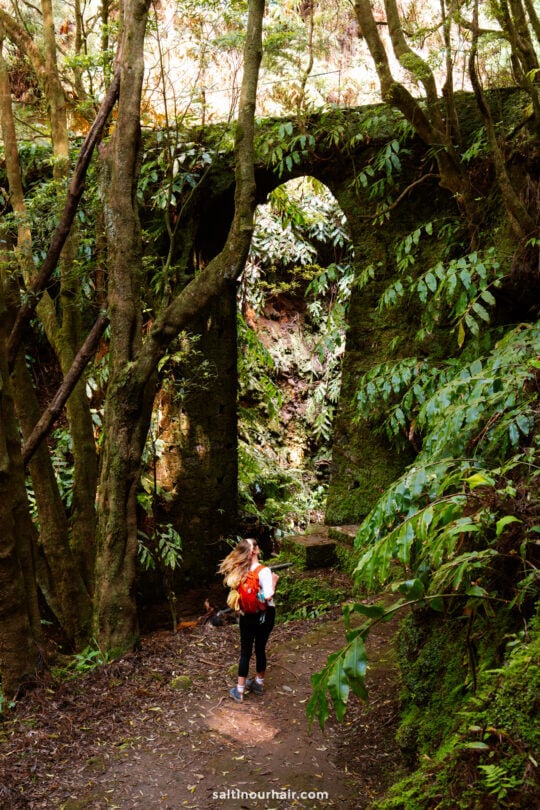A person with a red backpack stands on a forest path near a large, moss-covered stone arch surrounded by dense greenery—one of the unforgettable things to do in São Miguel for nature lovers and adventurers alike.