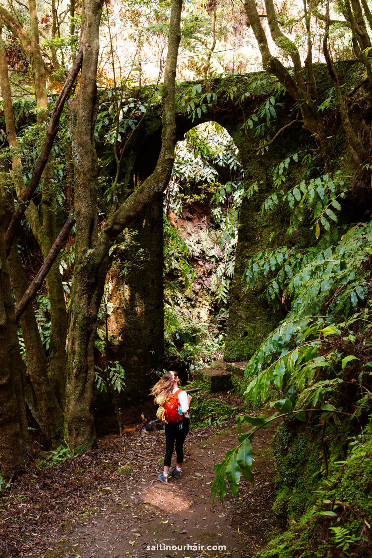 A woman with a red backpack stands on a forest path near a large, moss-covered stone arch surrounded by dense greenery on the PRC37 Janela do Inferno Hikeâ€”one of the unforgettable things to do in SÃ£o Miguel, Azores for nature lovers and adventurers.