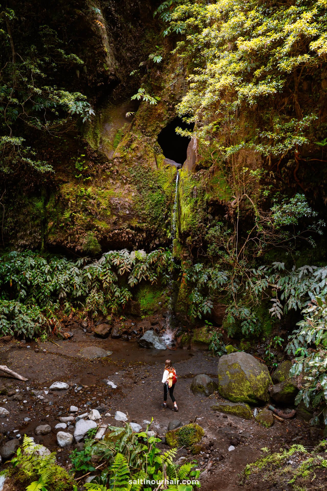 A woman with a backpack stands on rocky ground near  Janela do Inferno Waterfall flowing from a circular hole in a moss-covered cliff, surrounded by dense green foliageâ€”an adventurous moment among the many things to do in SÃ£o Miguel, Azores.