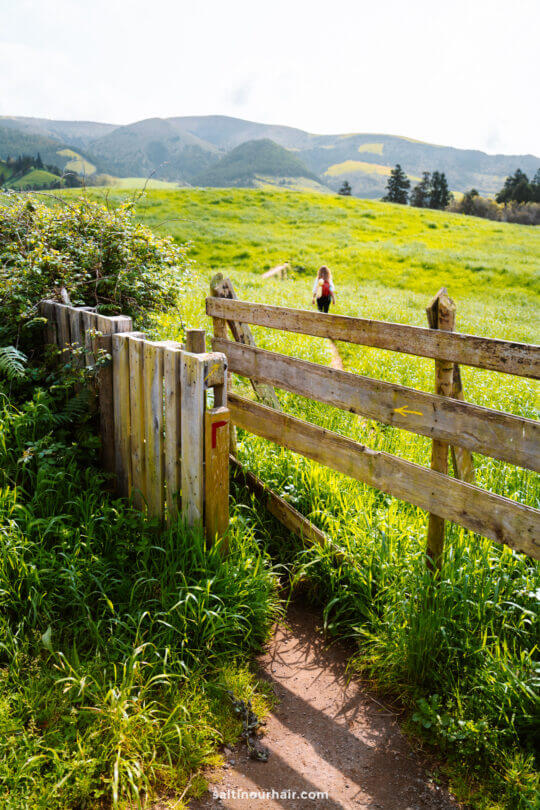 A wooden gate opens to a green field with a dirt path; a person with long hair walks away into the distance toward hills under a partly cloudy sky
