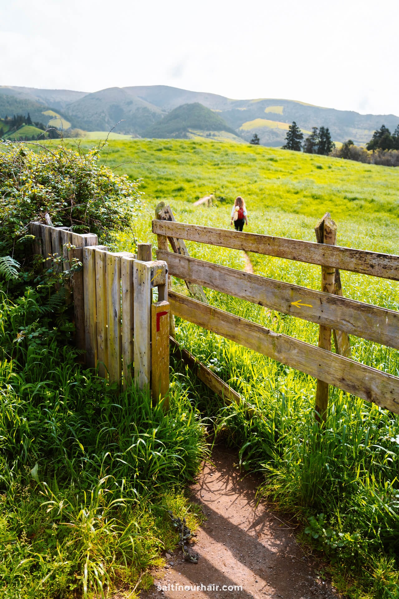 A wooden gate opens to a grassy field with a dirt path and a person walking in the distance; hills and trees are visible in the background, capturing the start of PRC37 Janela do Inferno, one of the best hikes in São Miguel, Azores.