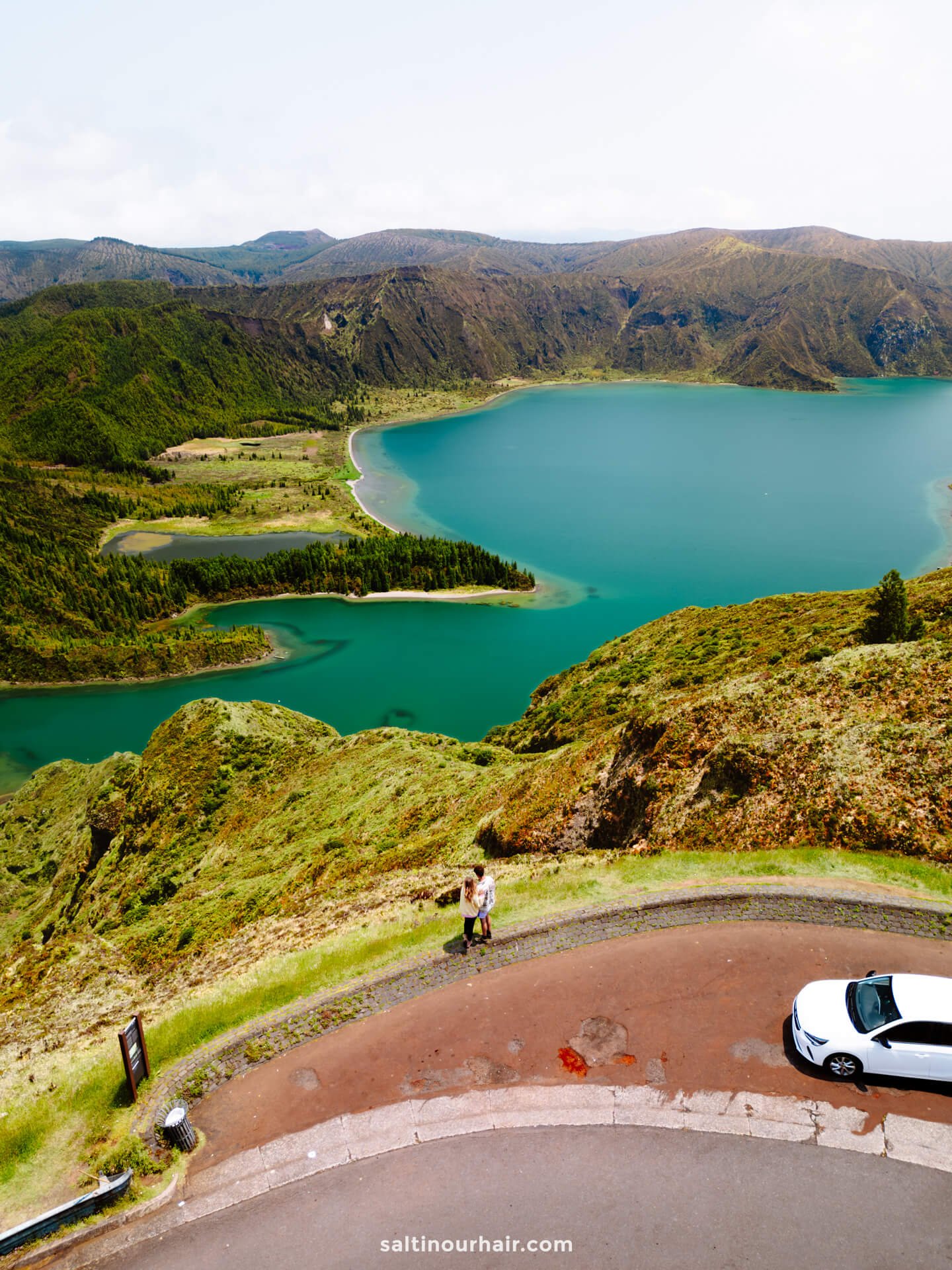 A couple stands on a curved road overlooking a turquoise lake and green mountains called Lagoa do Fogo, with a white car parked nearbyâ€”capturing one of the many unforgettable things to do in SÃ£o Miguel, Azores