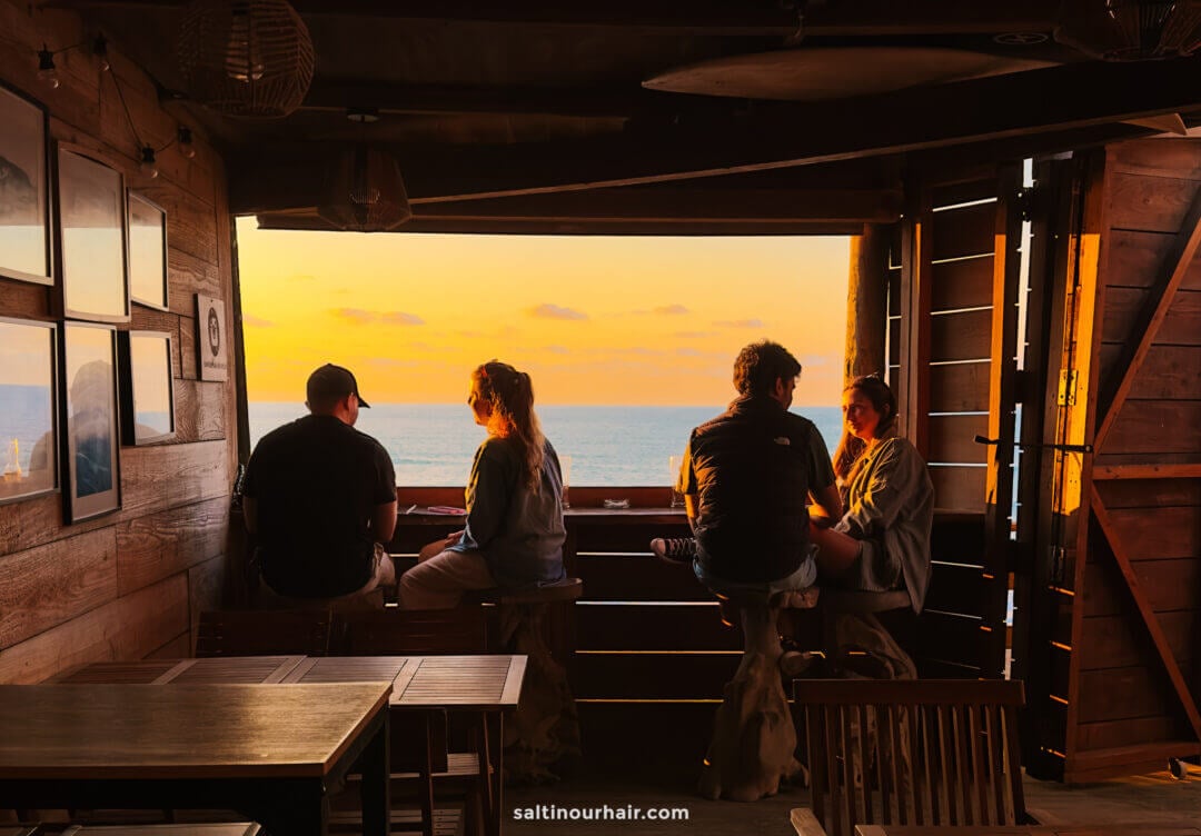 Four people sit on stools at a wooden bar in a cozy beach restaurant, facing an ocean sunset. Warm light fills the space, evoking the relaxed charm found in the Azores vs Madeira coastal retreats.