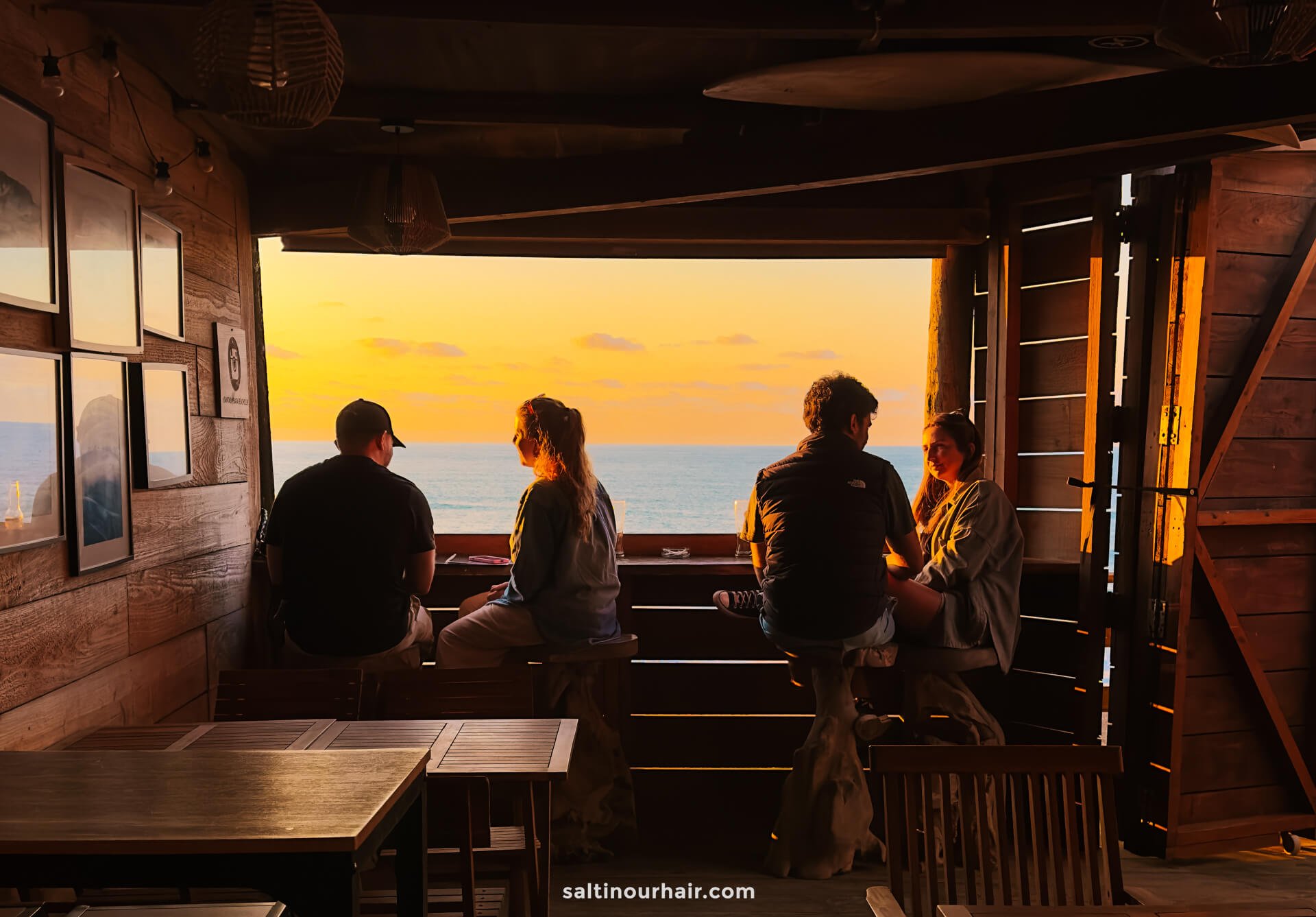 Four people sit at a wooden counter inside a rustic cabin, looking out at the ocean and sunset through an open windowâ€”a perfect way to unwind after exploring things to do in SÃ£o Miguel, Azores.