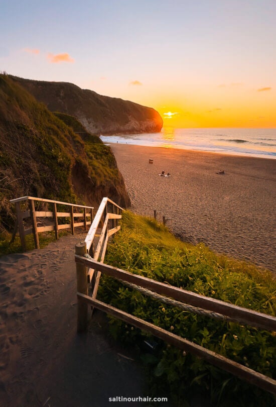 A sandy pathway with wooden railings leads down to Santa BÃ¡rbara Beach at sunset, with cliffs on one side and a few people sitting on the shoreâ€”a perfect spot for those seeking unique things to do in SÃ£o Miguel, Azores.