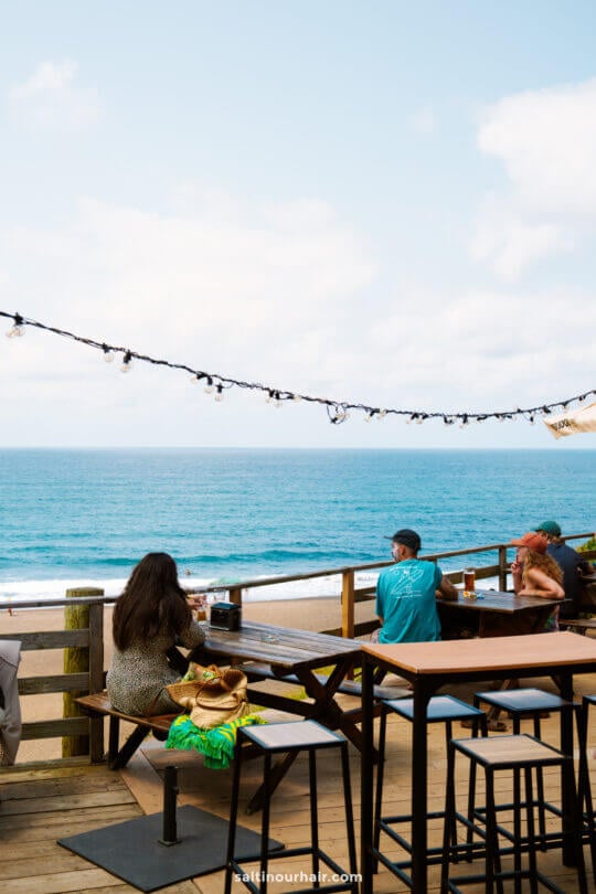 People sit at wooden tables on a deck overlooking the ocean under a partly cloudy sky with string lights aboveâ€”a perfect spot to unwind and enjoy one of the best things to do in SÃ£o Miguel, Azores.