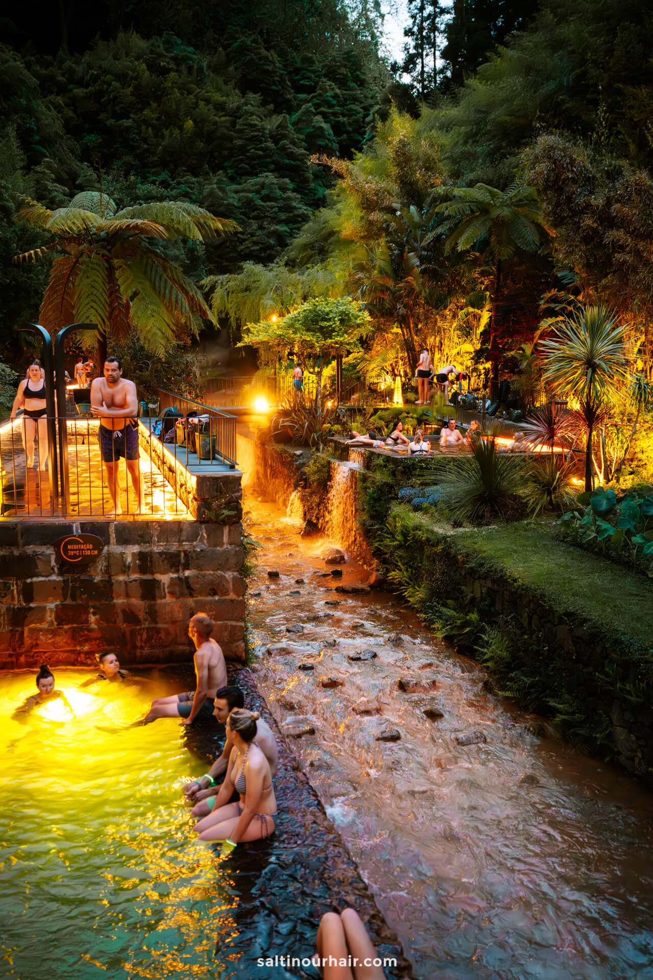 People relax in illuminated outdoor thermal pools surrounded by lush greenery and trees in a natural, tropical setting at duskâ€”one of the must-try things to do in SÃ£o Miguel, Azores.