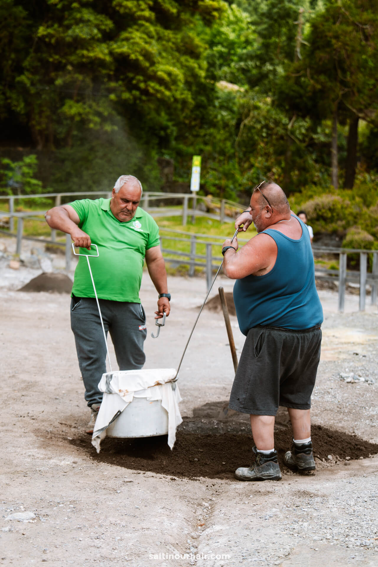 Two men stand on dirt, lifting a large pot out of the ground using metal rodsâ€”a traditional scene among the trees and wooden fence. Discovering this is one of the unique things to do in SÃ£o Miguel, Azores.