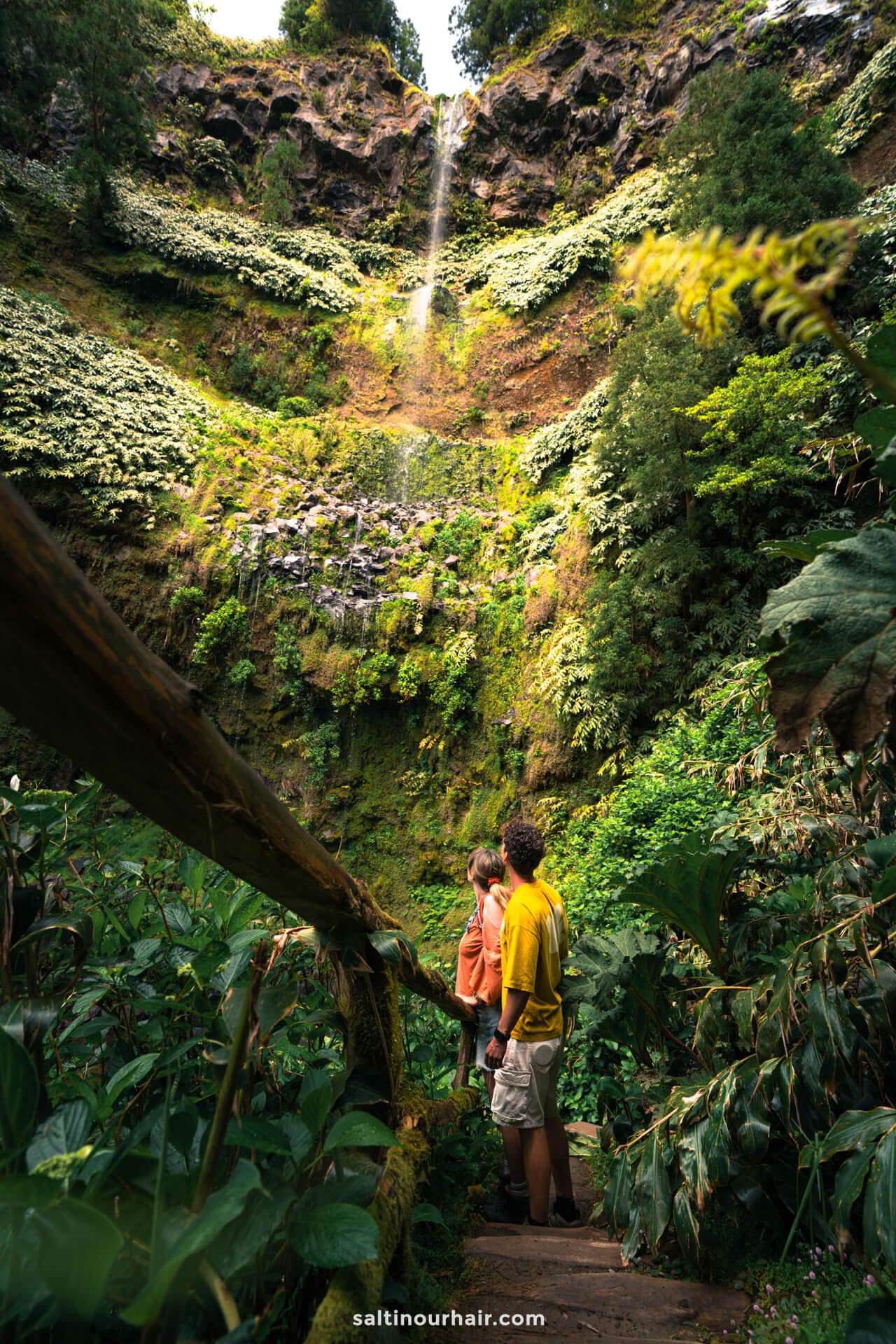 A young couple stand on a wooden path in Parque da GrenÃ¡ surrounded by dense green foliage, gazing up at a tall, thin waterfall cascading down a lush, rocky cliffâ€”one of the unforgettable things to do in SÃ£o Miguel, Azores.