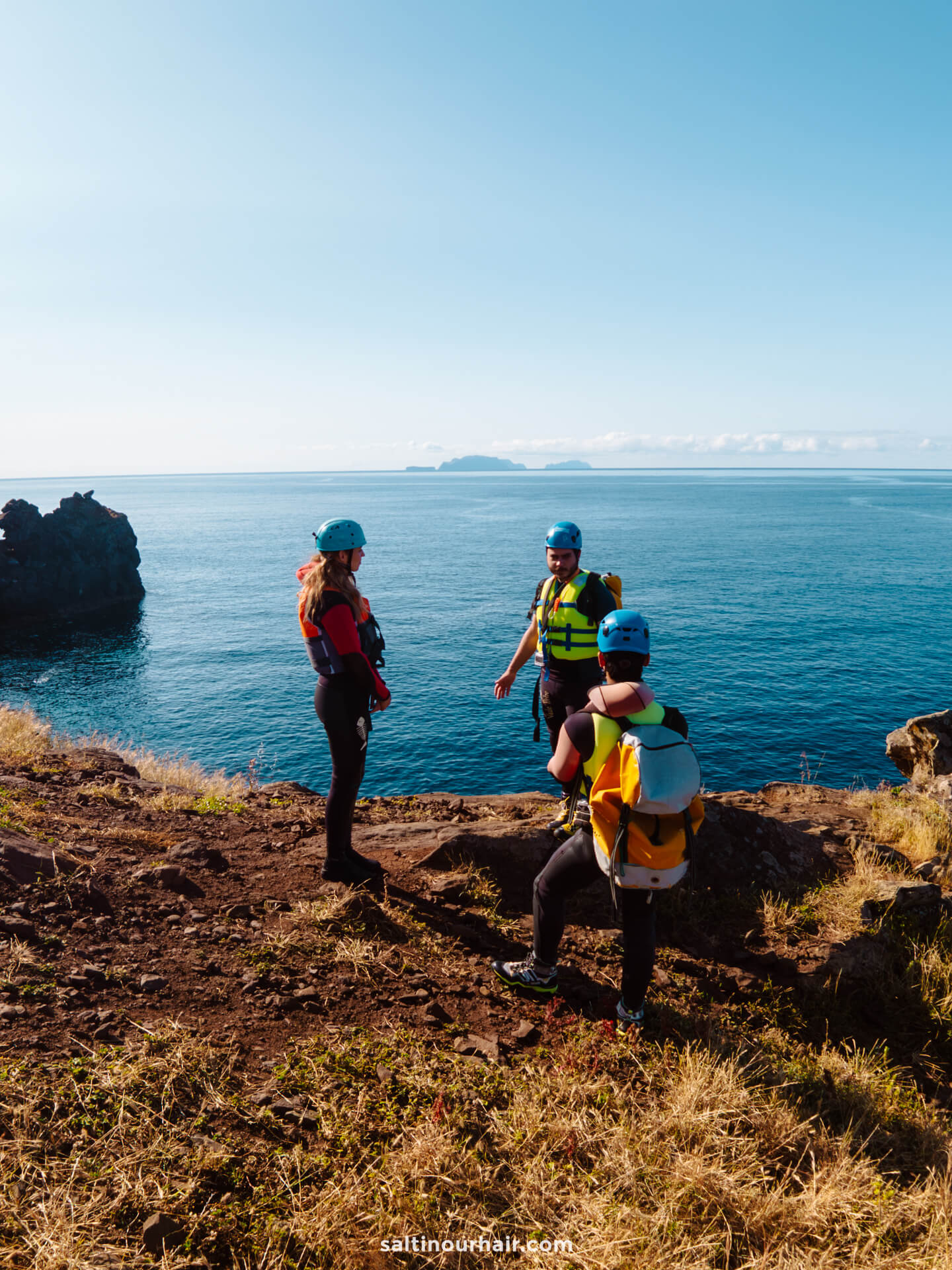 Three people in helmets and wetsuits stand on a rocky coastal cliff, ready for coasteering Madeira adventures, overlooking the ocean under a clear sky.