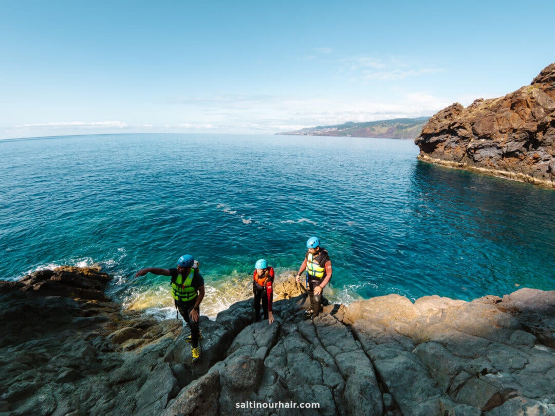 Three people in helmets and life jackets enjoy coasteering Madeira, climbing rocky cliffs beside a clear blue sea under a bright sky.