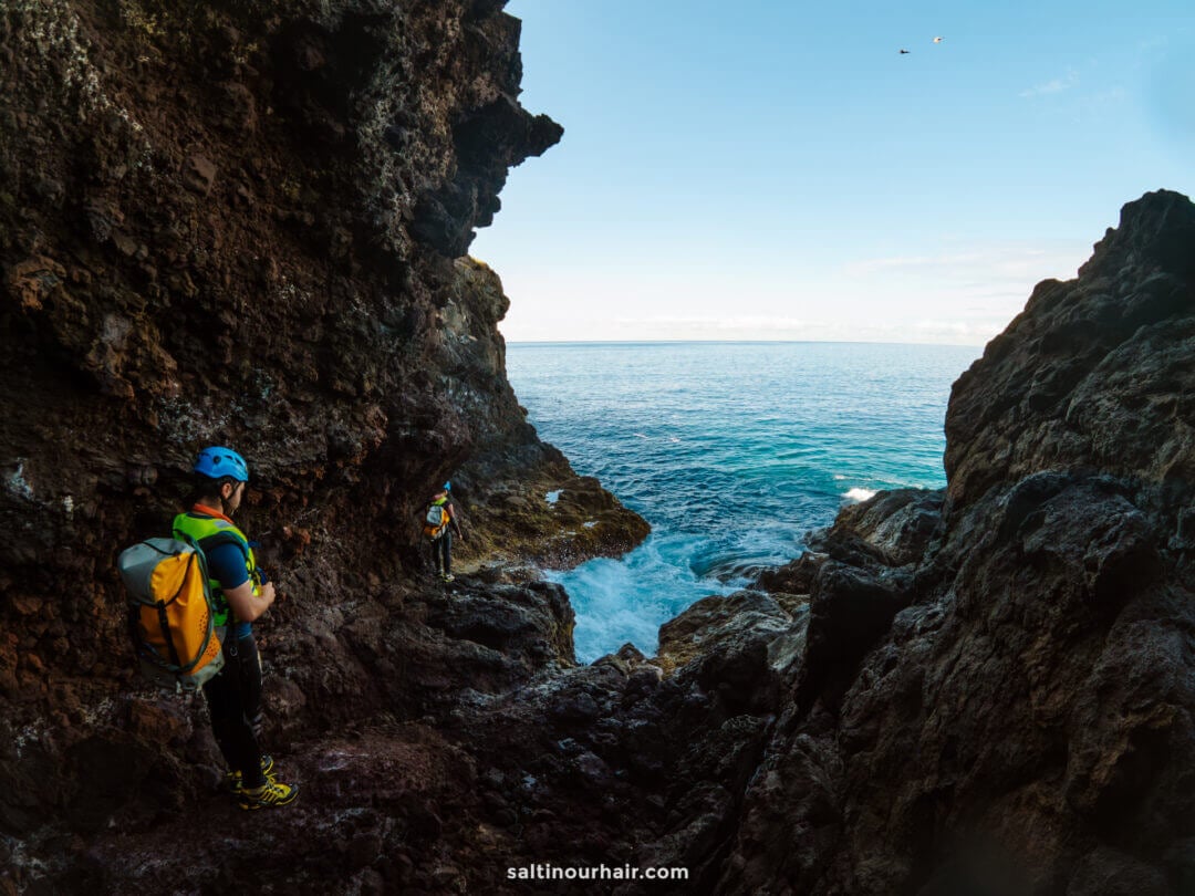 Two people wearing helmets and backpacks stand on rocky cliffs by the ocean, embodying the spirit of coasteering Madeira as they look out towards the water under a clear sky.