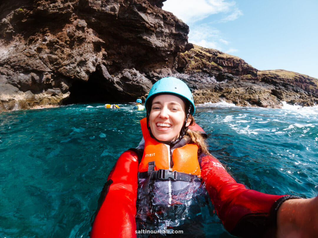 A woman in a red wetsuit, orange life jacket, and blue helmet takes a selfie while coasteering Madeira, floating in blue water near rocky cliffs and a cave.