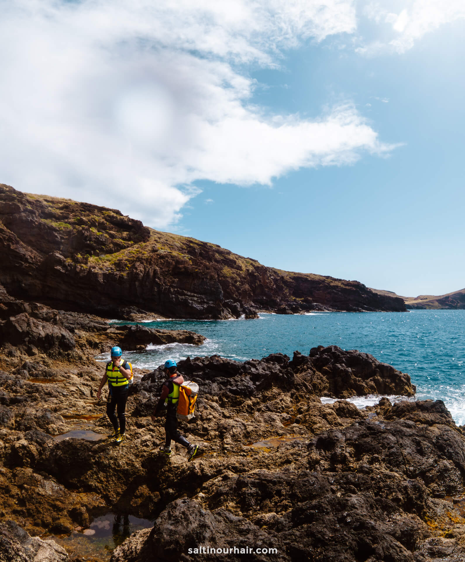 Two people in wetsuits and helmets walk on rocky coastal terrain near the ocean under a partly cloudy sky, experiencing the thrill of coasteering Madeira.