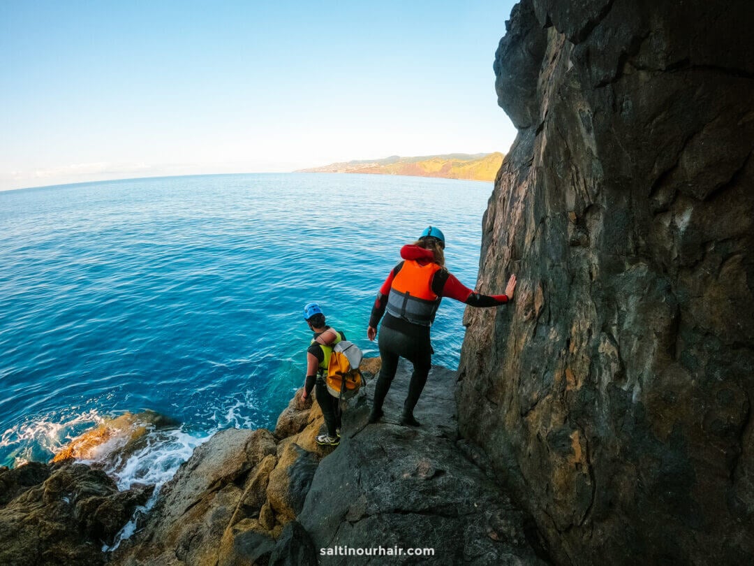 Two people wearing wetsuits and life jackets walk along a rocky cliff edge by the ocean under a clear sky, enjoying an adventurous coasteering Madeira experience.