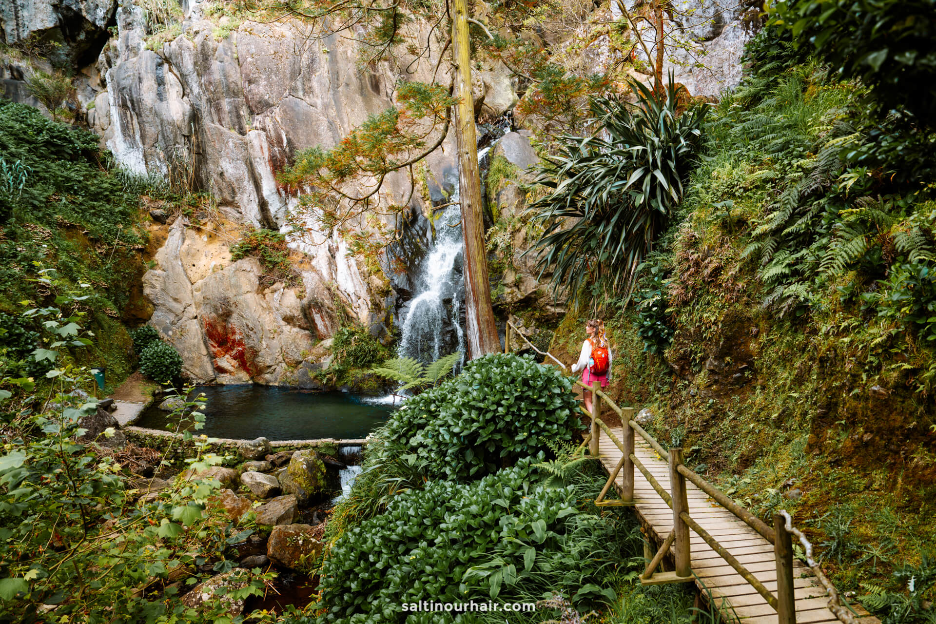 A person stands on a wooden bridge surrounded by dense greenery, facing a small waterfall flowing into a pool at the base of rocky cliffsâ€” Cascata do Limbo is the perfect spot for those seeking adventures things to do in SÃ£o Miguel, Azores.