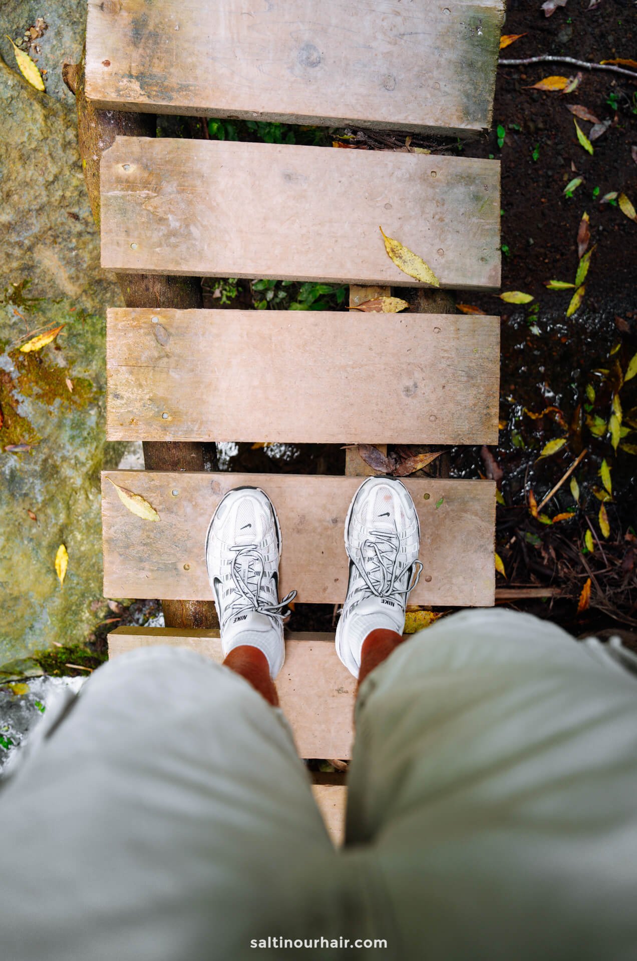 Man wearing white sneakers and light shorts standing on a wooden plank bridge, viewed from above, with leaves and ground visible below