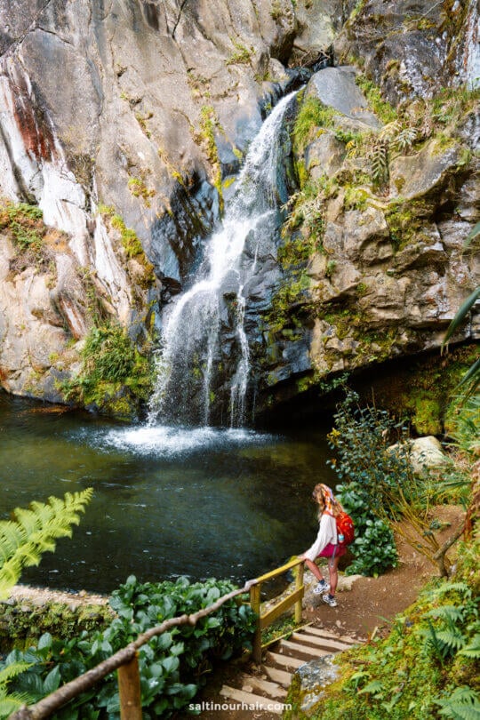A person with a red backpack stands on a wooden path next to a small waterfall cascading into a rocky pool surrounded by greenery, capturing one of the must-see things to do in São Miguel.