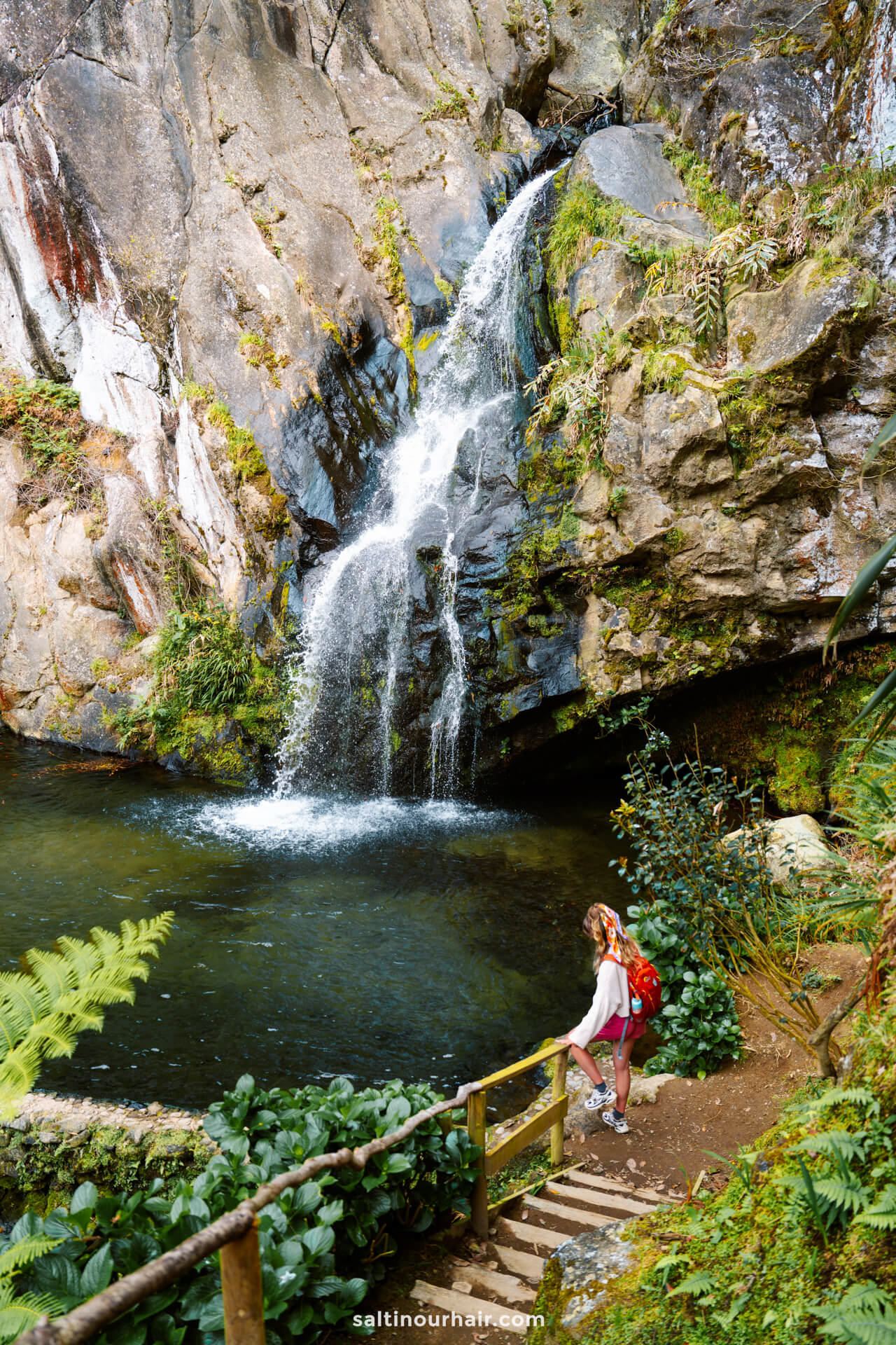 A woman with a red backpack stands on a wooden path next to a small waterfall called Cascata do Limbo cascading into a rocky pool surrounded by greenery, capturing one of the must-see things to do in SÃ£o Miguel.