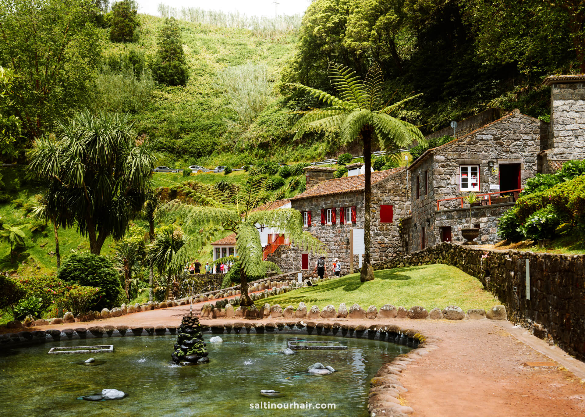 A stone house with red shutters sits beside a pond and lush greenery at Parque Natural da Ribeira dos CaldeirÃµes, where people walk along a path in this hilly, forested areaâ€”one of the scenic things to do in SÃ£o Miguel, Azores.