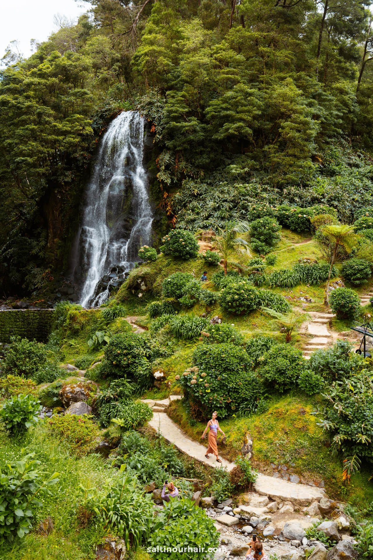 A tall waterfall flows down a cliff surrounded by dense green foliage at Parque Natural da Ribeira dos CaldeirÃµes, with people walking along a winding stone path belowâ€”a must-see among the top things to do in SÃ£o Miguel, Azores.