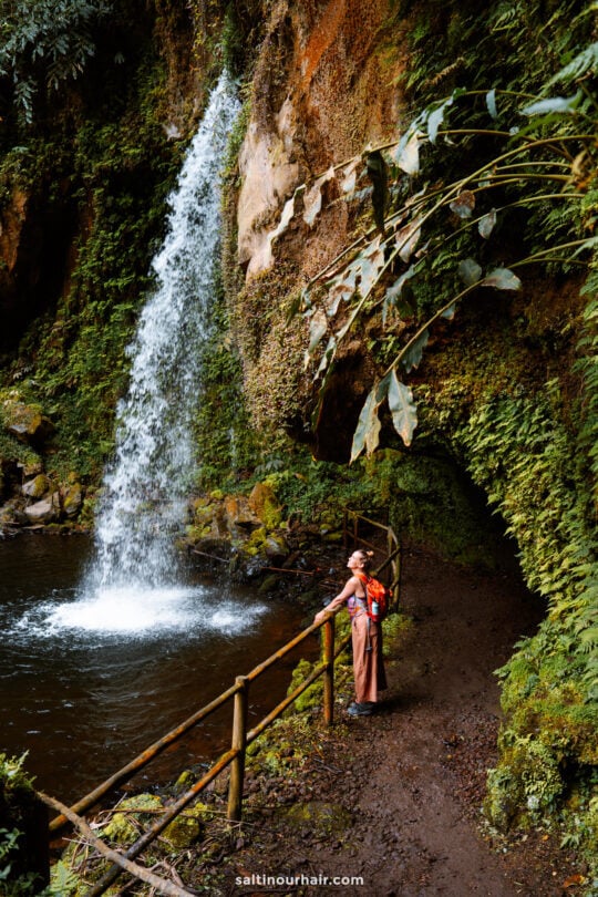 A person with a red backpack stands on a muddy trail by a railing, looking at a waterfall cascading into a pool surrounded by lush greeneryâ€”one of the unforgettable things to do in SÃ£o Miguel.