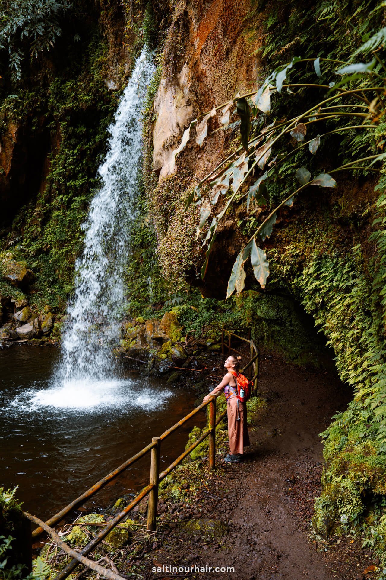 A woman with a red backpack stands on a muddy trail by a railing, looking at a waterfall cascading into a pool surrounded by lush greeneryâ€”Cascata da Gruta is one of the unforgettable things to do in SÃ£o Miguel, Azores