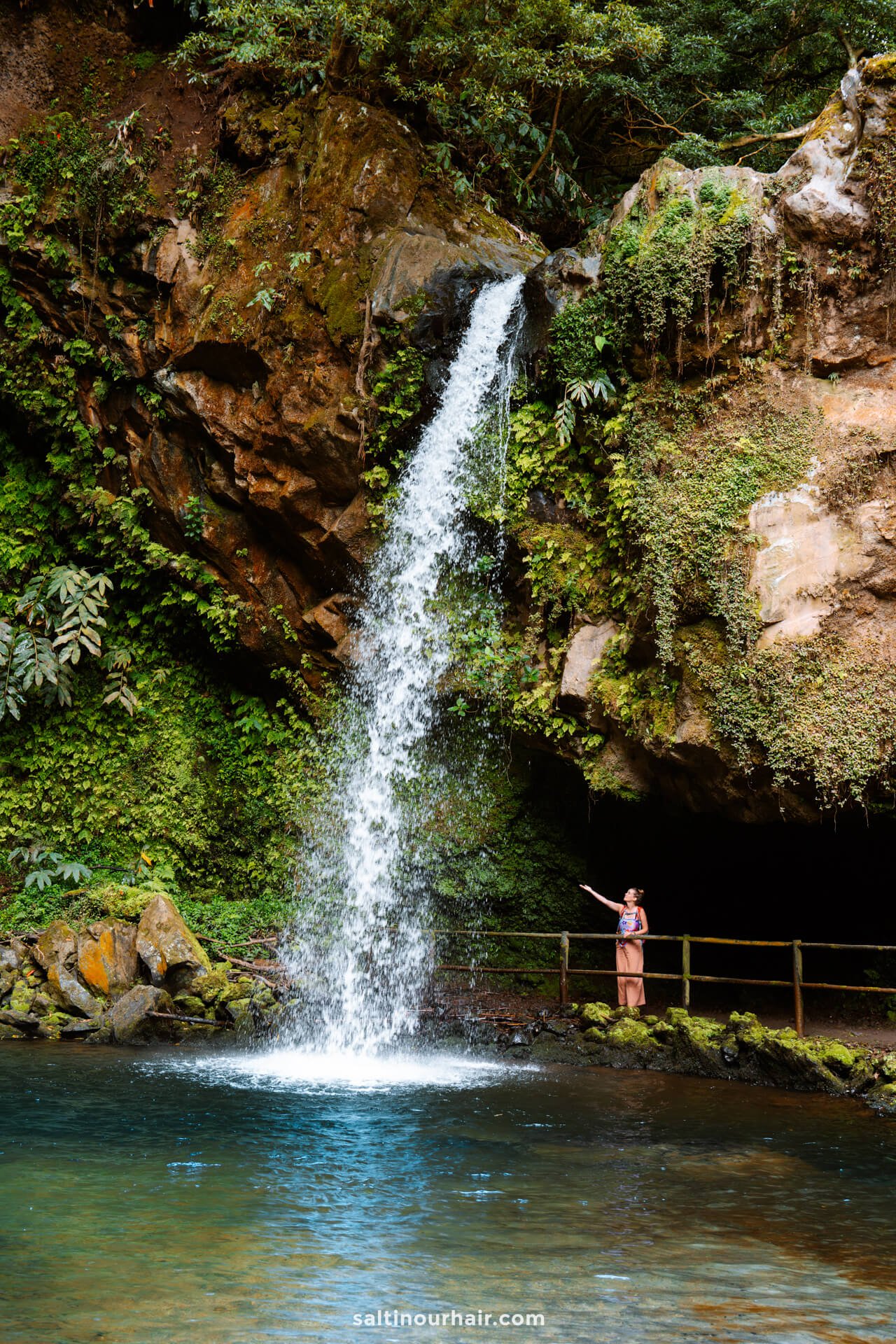 A woman stands on a railing next to a small waterfall called Cascata da Gruta, flowing from a rocky cliff surrounded by lush greenery into a clear poolâ€”one of the enchanting things to do in SÃ£o Miguel, Azores.