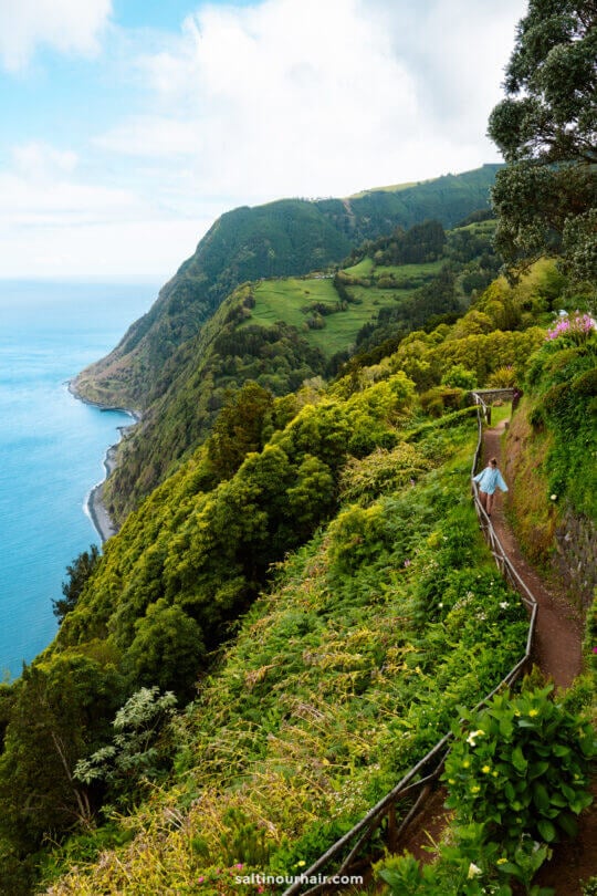 A person walks along a narrow path surrounded by lush green cliffs overlooking the ocean, with a coastline and hills in the backgroundâ€”a perfect scene among the top things to do in SÃ£o Miguel.