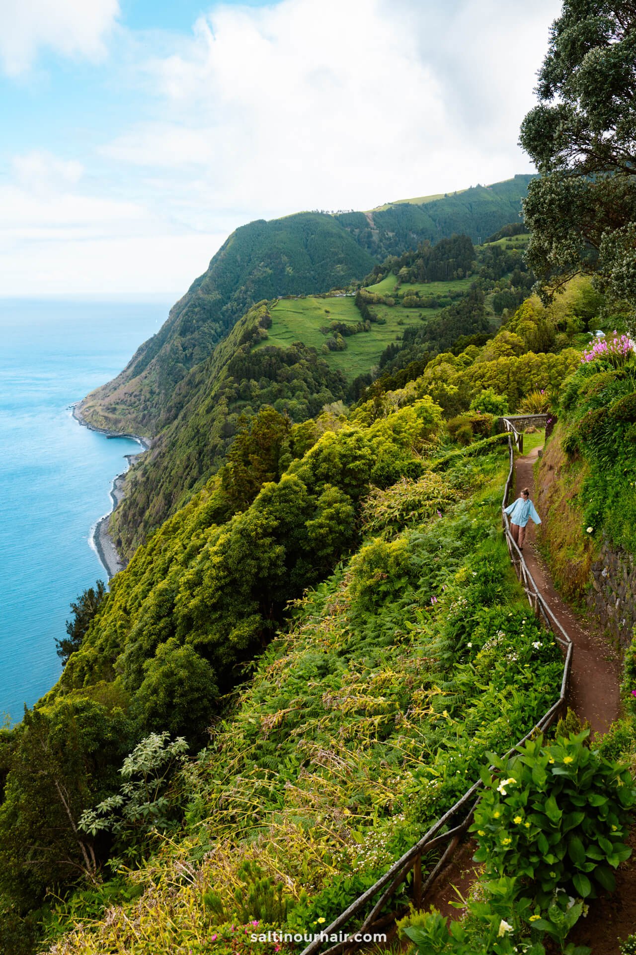 A woman walks along a narrow path surrounded by lush green cliffs overlooking the ocean, with a coastline and hills in the background at Miradouro da Ponta do Sossegoâ€”a perfect scene among the top things to do in SÃ£o Miguel, Azores.