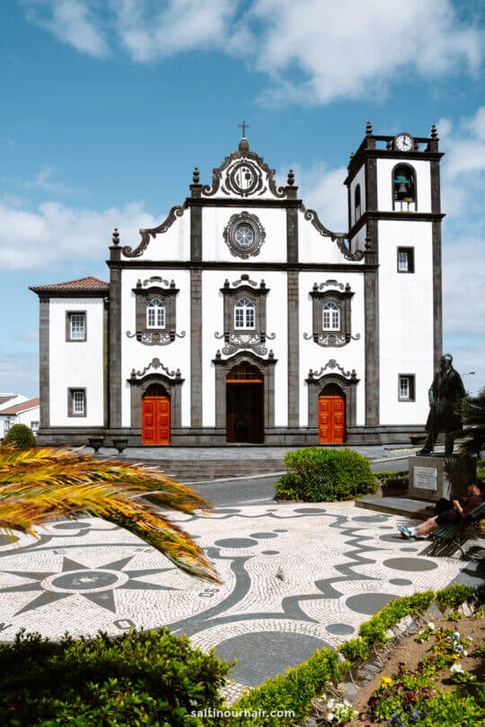 A white and black baroque-style church with red doors, a bell tower, and a patterned stone plaza in the foreground under a partly cloudy sky—one of the top things to do in São Miguel.