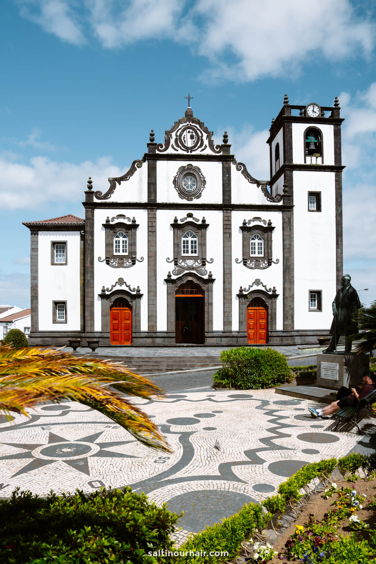 A white and black baroque-style church with red doors, a bell tower, and a patterned stone plaza in the foreground in Nordeste, SÃ£o Miguel Azores.