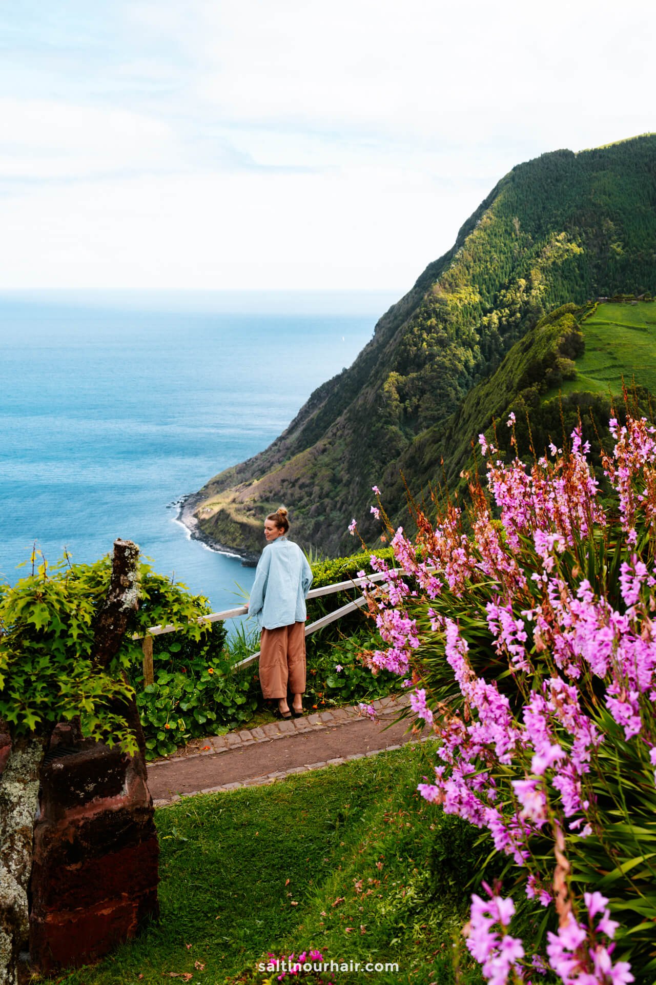 A woman stands on a path overlooking a coastal cliff at Miradouro da Ponta do Sossego, with pink flowers in the foreground and the ocean and green hills in the backgroundâ€”one of the breathtaking things to do in SÃ£o Miguel, Azores.