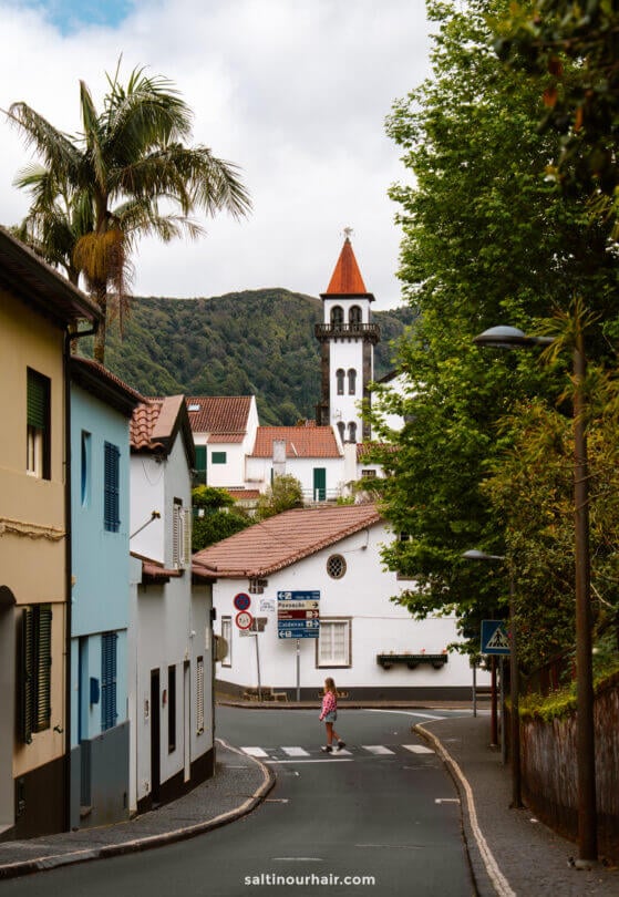 A woman crossed a narrow street with colorful houses in the small town of Frunas, with a church steeple and green hills visible in the backgroundâ€”capturing the charm of everyday life and inspiring ideas for things to do in SÃ£o Miguel, Azores.