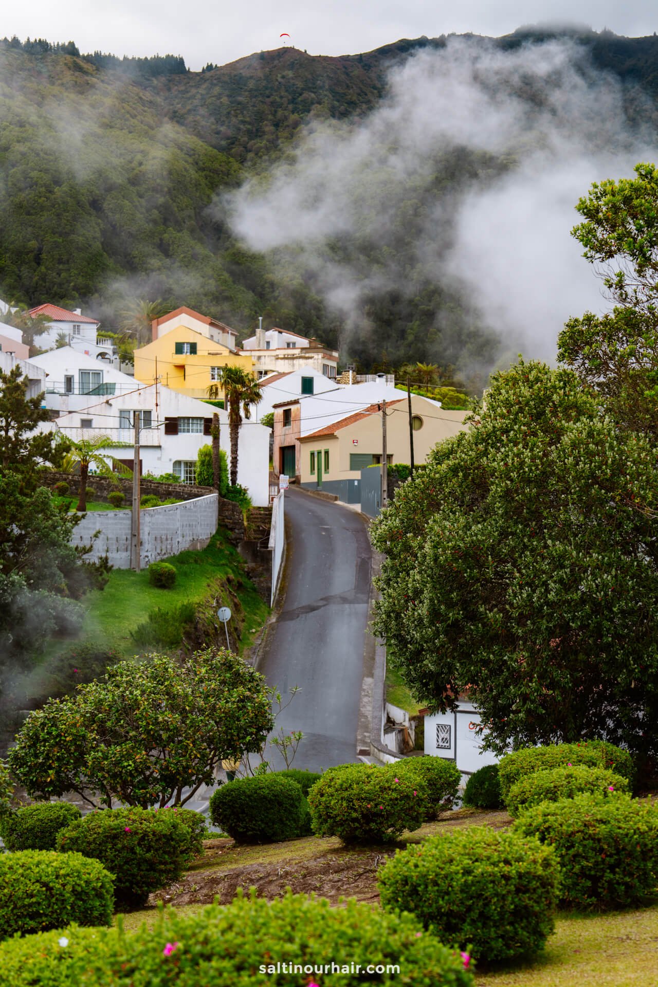 A narrow road leads through a lush, green village with white and yellow houses, surrounded by mist and hills—one of the locations where to stay in Furnas sao miguel azores