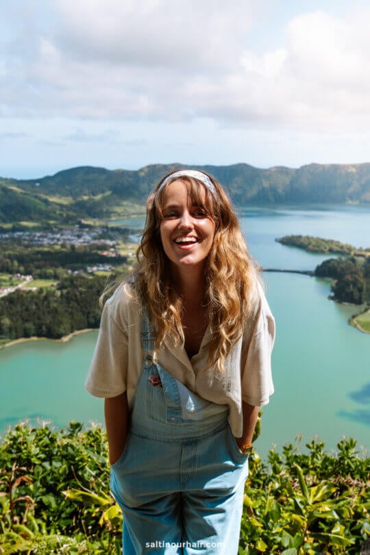 A young woman wearing a white shirt and denim overalls stands in front of a scenic lake and mountain landscape at Sete Cidades, smiling at the cameraâ€”one of the many unforgettable things to do in SÃ£o Miguel, Azores.