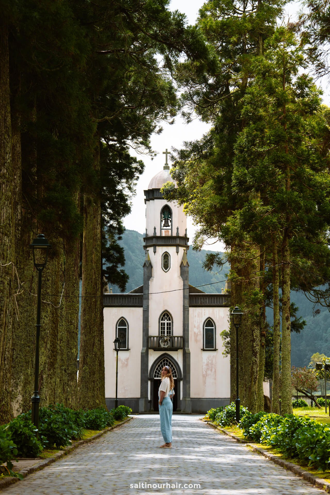 A woman in a light blue trousers stands on a tree-lined path facing a white church with a bell tower and cross in Sete Cidades, capturing one of the must-see things to do in SÃ£o Miguel, Azores.
