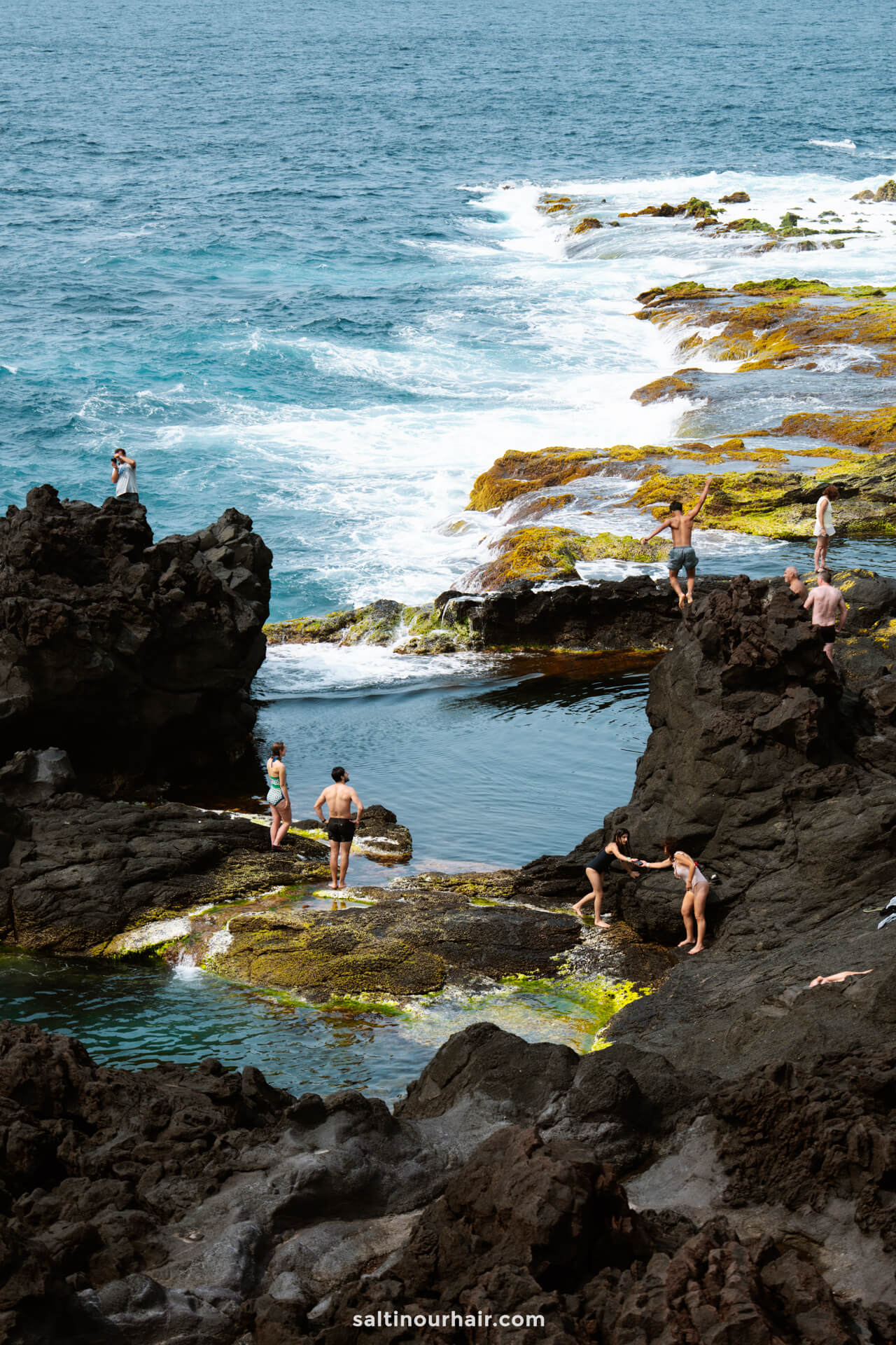 People swim, climb, and relax on rugged black volcanic rocks and tide pools beside the ocean, with waves crashing on the mossy shorelineâ€”one of the unforgettable things to do in SÃ£o Miguel, Azores.
