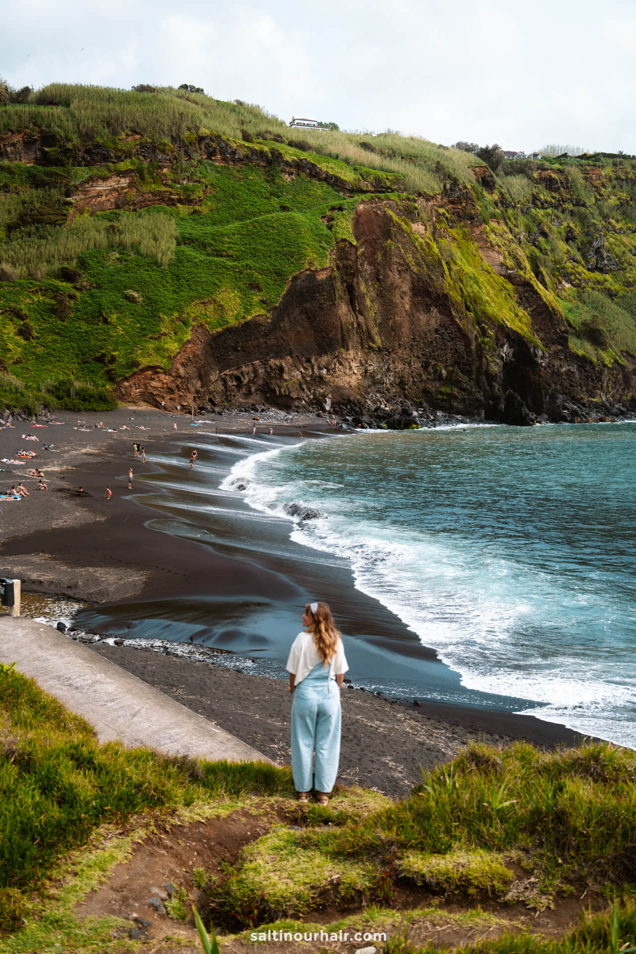 A woman stands on a grassy cliff overlooking a black sand beach called: Praia dos Mosteiros, with people sunbathing and swimming, steep green hills in the backgroundâ€”a scenic spot among the top things to do in SÃ£o Miguel, Azores