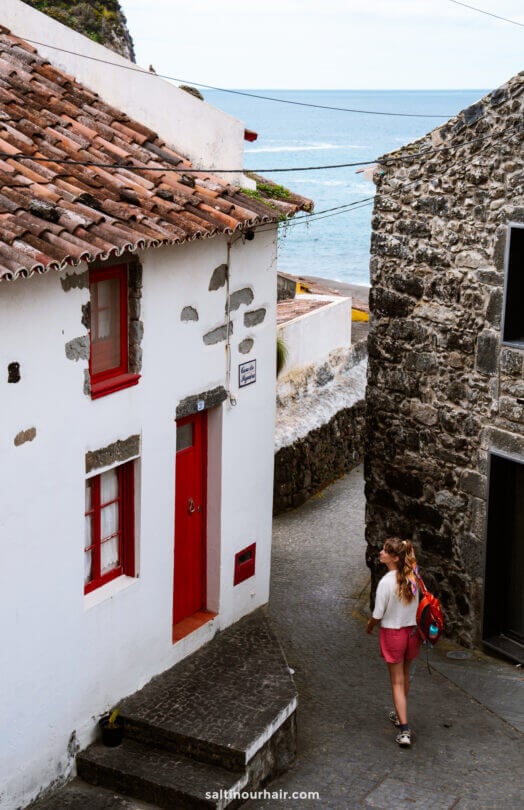 A woman with a backpack walks down a narrow cobblestone street lined with stone buildings near the ocean, exploring one of the charming things to do in SÃ£o Miguel, Azores.