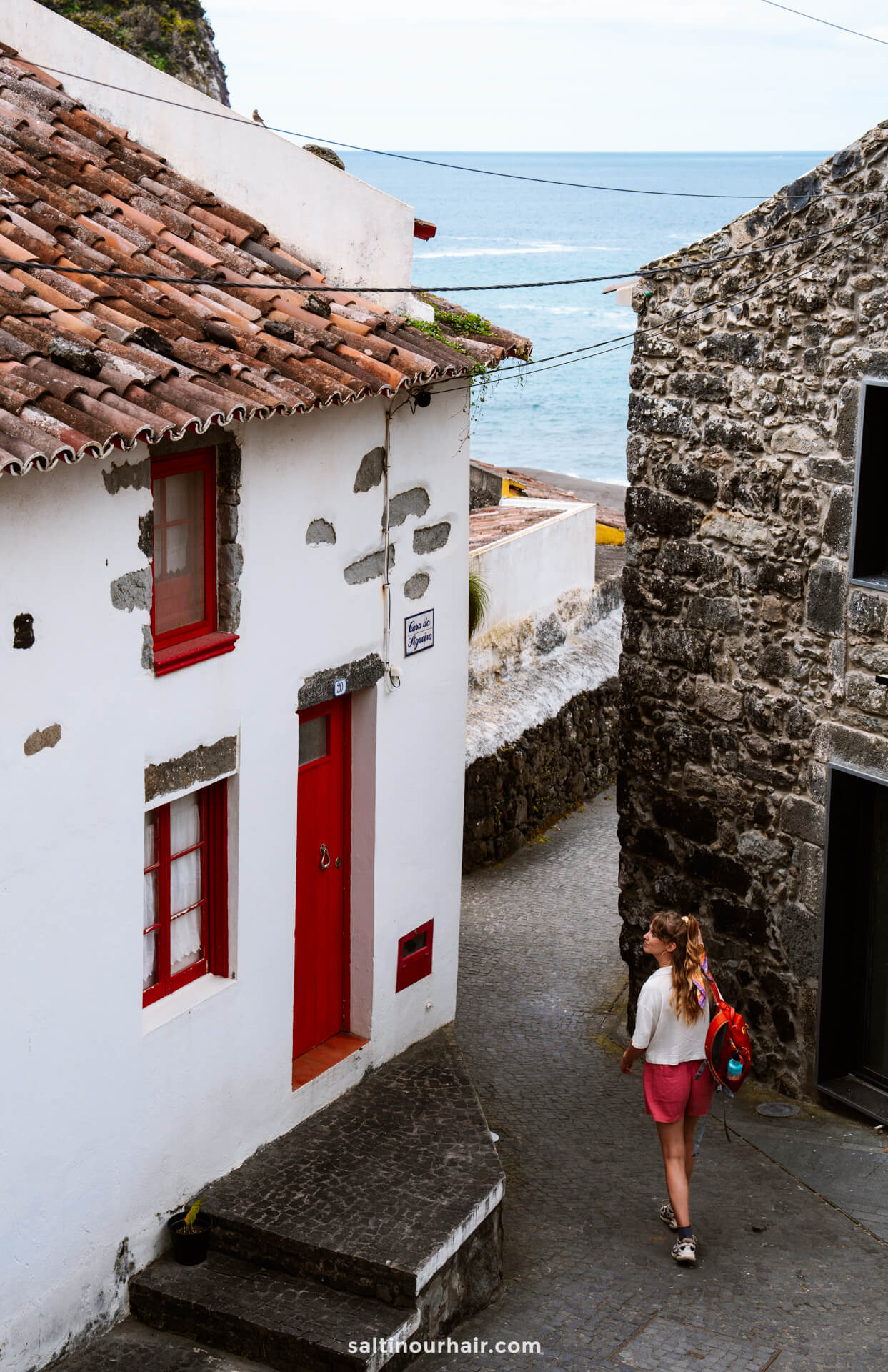 A woman walks down a narrow cobblestone street lined with stone buildings near the ocean, discovering where to stay near the beaches in sao miguel azores