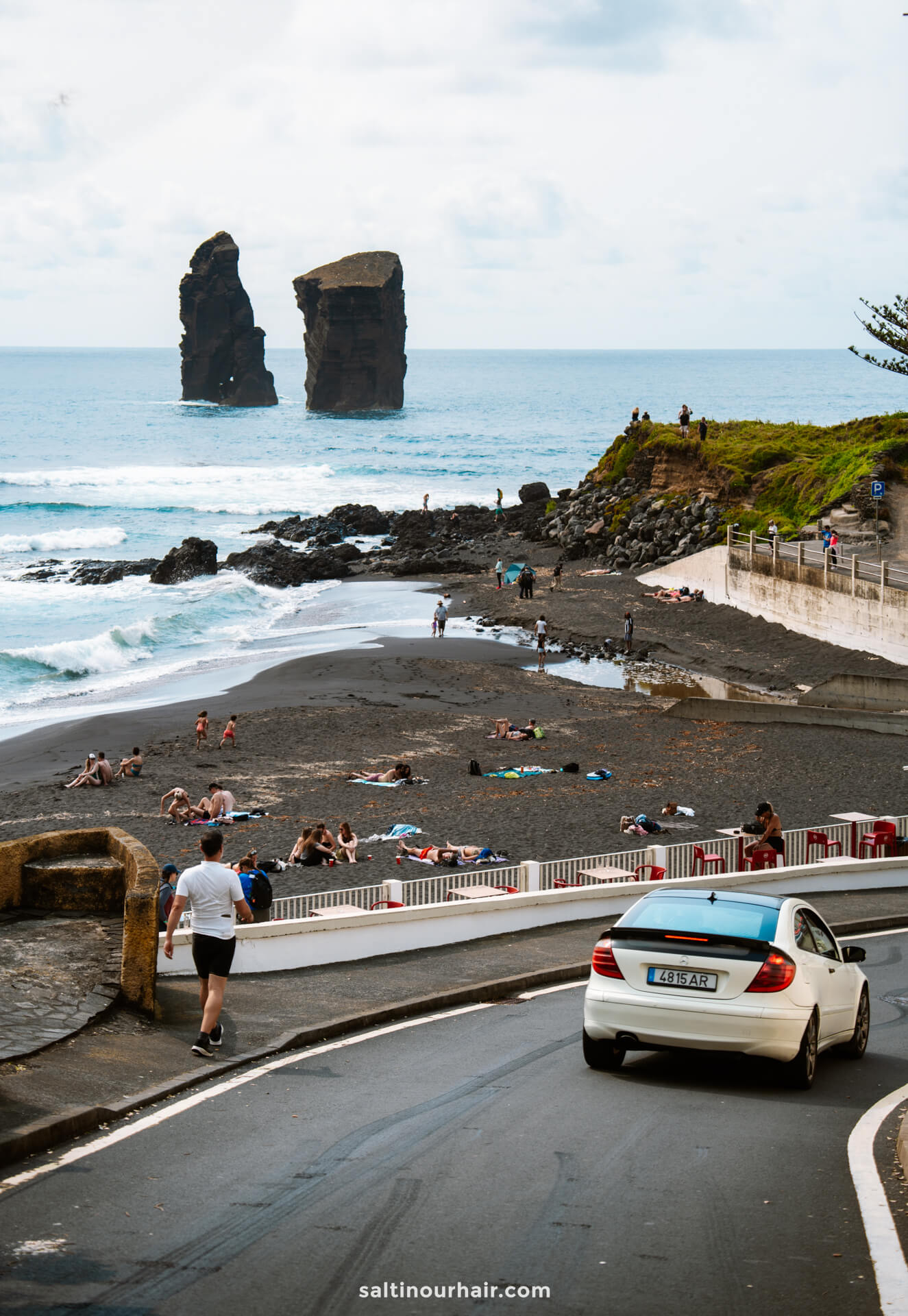 People relax on a black sand beach near the ocean, with two large rock formations in the waterâ€”a must-see among things to do in SÃ£o Miguel. A car drives down a road in the foreground.