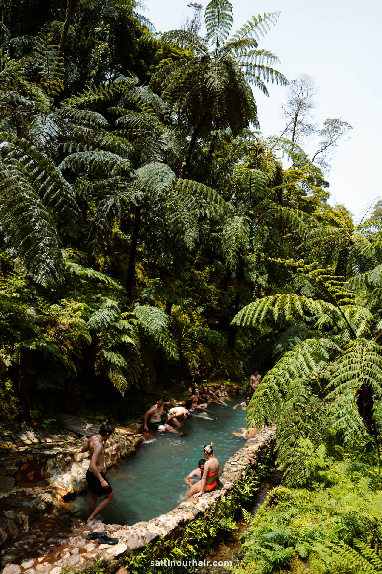 A group of people relax in a narrow, natural hot spring pool surrounded by dense, green tropical vegetation and tall fernsâ€”one of the unforgettable things to do in SÃ£o Miguel, Azores.