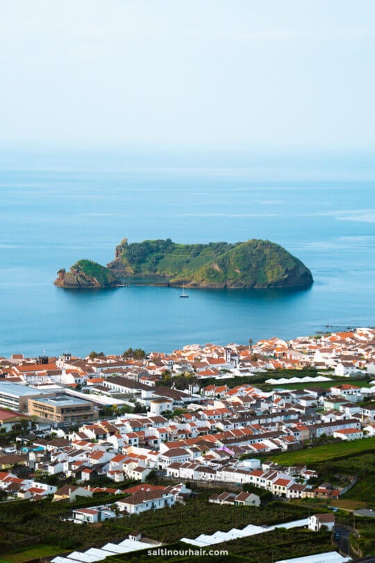 A small green island sits in the blue ocean near a coastal town of Vila FrancaÂ with white buildings and red roofs, offering a glimpse of the many things to do in SÃ£o Miguel, Azores