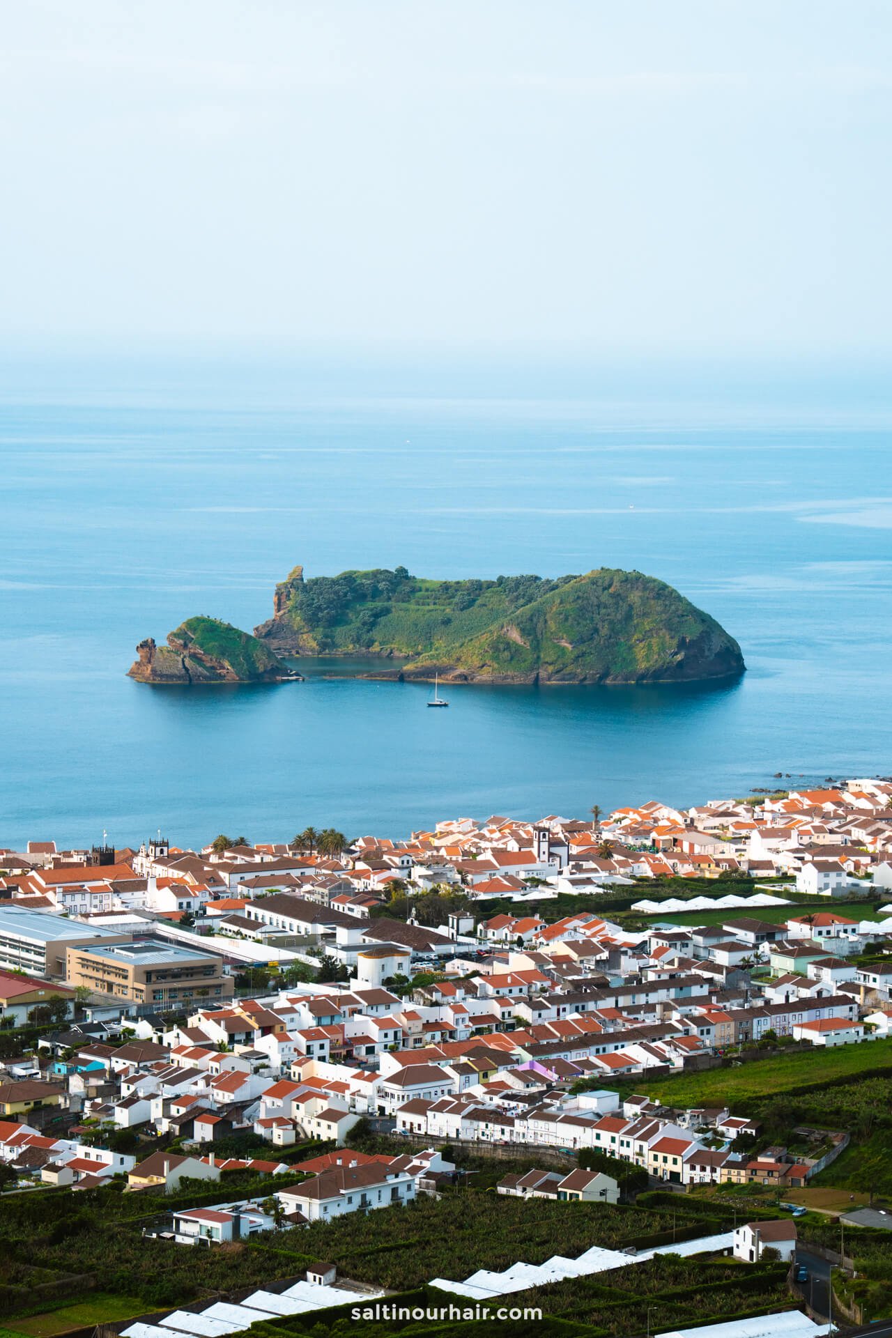 A small green island sits in the blue ocean near a coastal town with white buildings and red roofs, offering a glimpse of the many things to do in São Miguel.