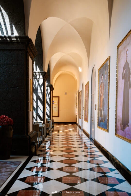 A hallway with arched ceilings, large portraits on the walls, geometric patterned floor tiles, and sunlight casting shadows through windowsâ€”capturing the charm you might find while exploring things to do in SÃ£o Miguel.