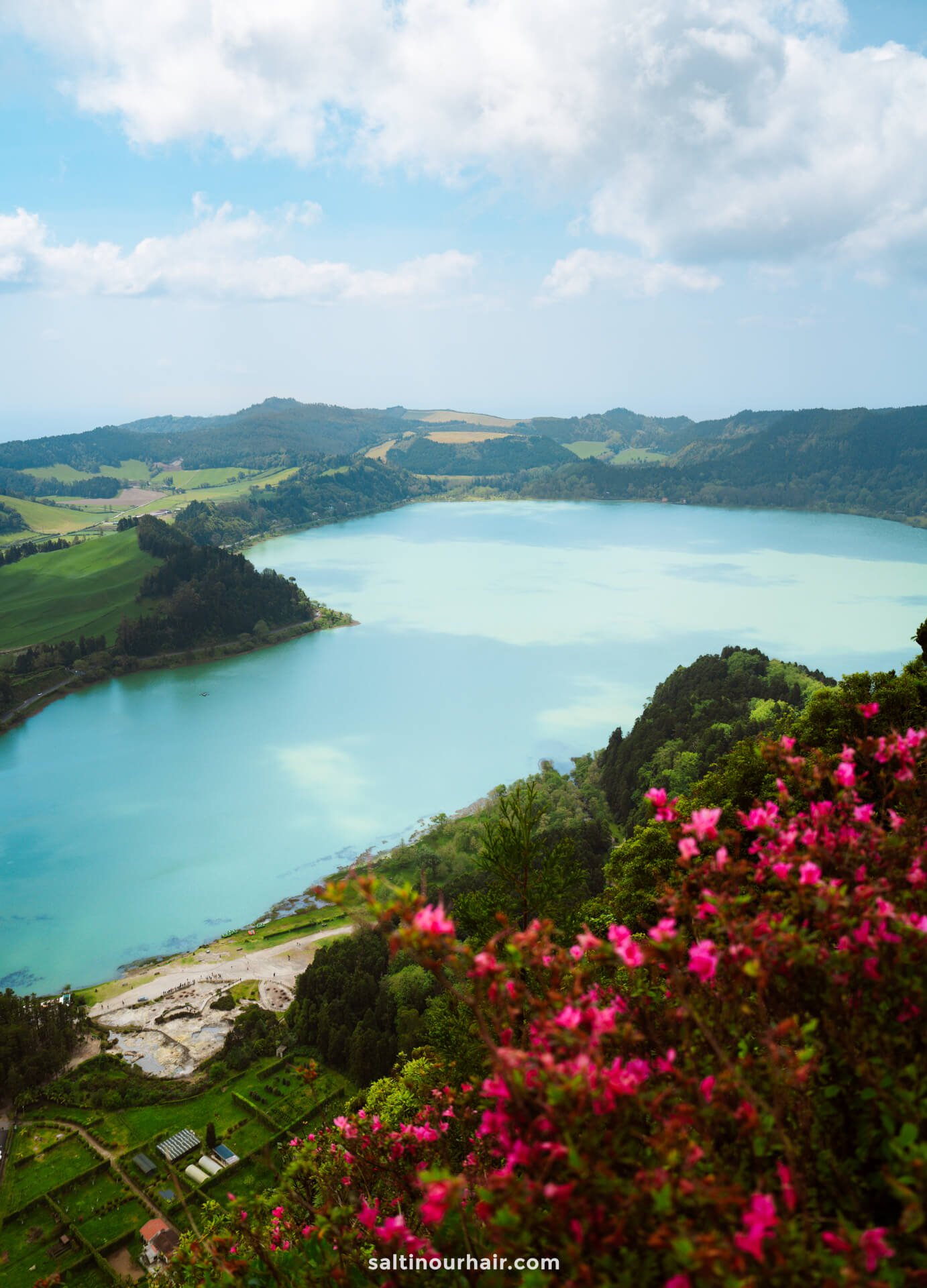 View of a turquoise lake called Lagoa das Furnas surrounded by green hills and fields, with pink flowers in the foreground under a partly cloudy skyâ€”a perfect spot to add to your list of things to do in SÃ£o Miguel, Azores.